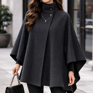 Puede incluir: Una capa gris carbón con cuello alto y cierre de botones. La capa tiene un diseño fluido y se combina con pantalones oscuros y un bolso negro con una correa de cadena dorada. La persona lleva gafas de sol y joyas de oro.