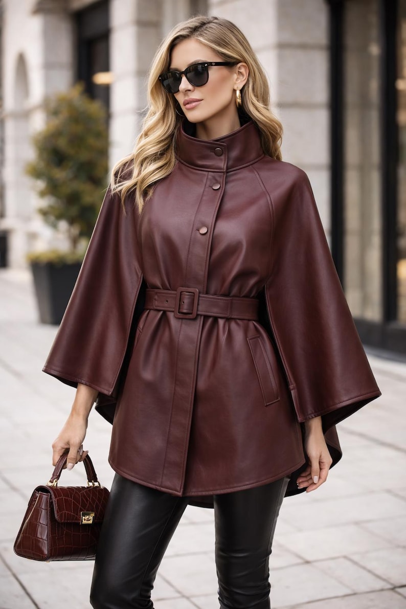 May include: A burgundy leather cape coat with a high collar and a matching belt. The coat has a button closure and flared sleeves. A matching burgundy handbag is held in the hand.