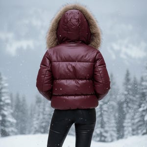 K&ouml;nnte beinhalten: Eine bordeauxfarbene Steppjacke mit Kapuze mit Pelzbesatz. Die Jacke wird mit einer schwarzen Hose getragen. Der Hintergrund zeigt einen verschneiten Wald. Die Jacke scheint aus einem gl&auml;nzenden Material zu sein.