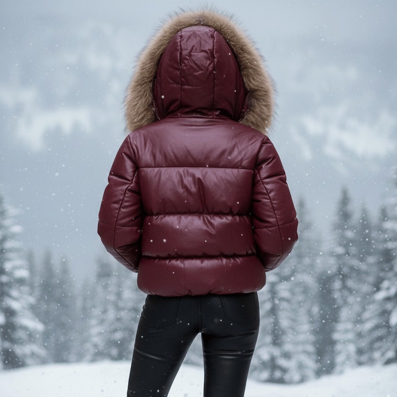 K&ouml;nnte beinhalten: Eine bordeauxfarbene Steppjacke mit Kapuze mit Pelzbesatz. Die Jacke wird mit einer schwarzen Hose getragen. Der Hintergrund zeigt einen verschneiten Wald. Die Jacke scheint aus einem gl&auml;nzenden Material zu sein.