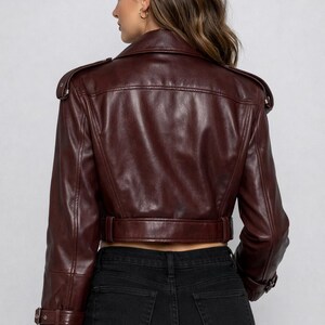 May include: A burgundy leather jacket with a cropped design. The jacket features a notched collar, shoulder epaulets, and a belt at the waist. The sleeves have buckle details. The model is wearing black jeans.