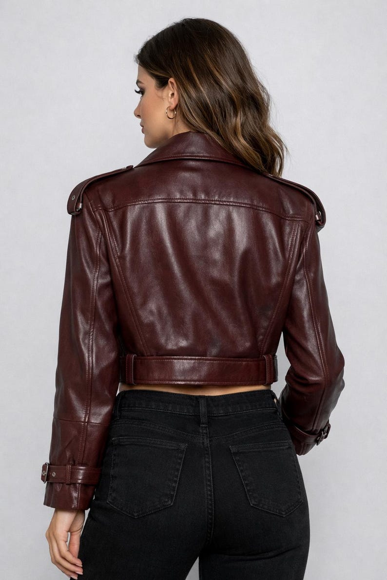 May include: A burgundy leather jacket with a cropped design. The jacket features a notched collar, shoulder epaulets, and a belt at the waist. The sleeves have buckle details. The model is wearing black jeans.