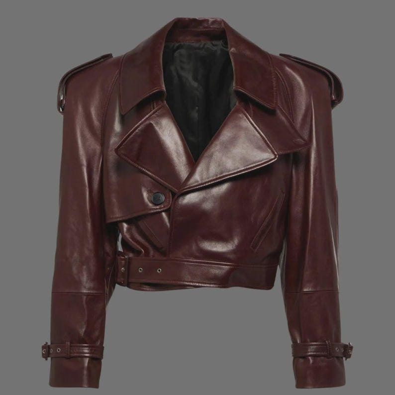 May include: A burgundy leather jacket with a classic trench coat design. The jacket features a notched collar, shoulder epaulets, a belted waist, and adjustable cuffs. The jacket has a single-button closure and a cropped length.