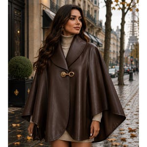 May include: A dark brown leather cape featuring a wide collar and a single gold-toned metal toggle closure at the neckline. The garment is worn over a beige turtleneck sweater, showcasing a smooth finish and a structured, flowing silhouette.