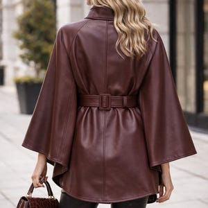 May include: A dark brown leather cape jacket with a matching belt cinching the waist. The jacket has wide sleeves and a high collar. A brown handbag with a crocodile pattern is held in the hand.