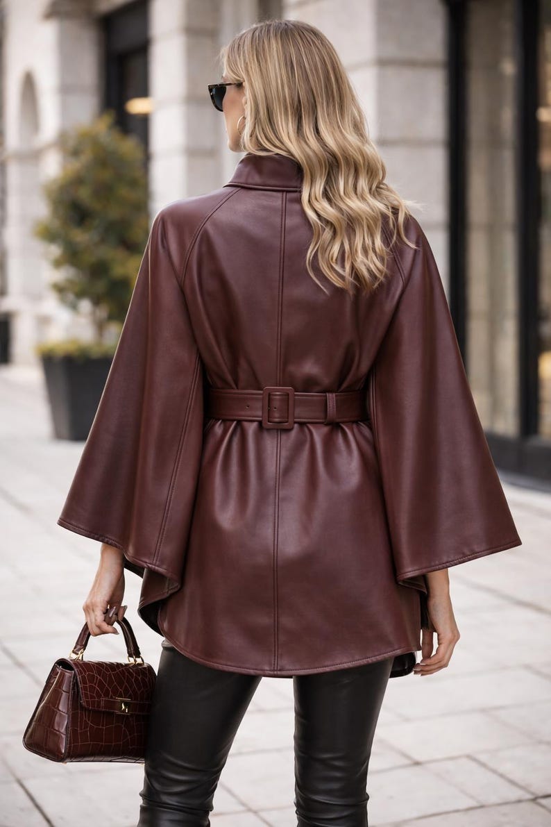 May include: A dark brown leather cape jacket with a matching belt cinching the waist. The jacket has wide sleeves and a high collar. A brown handbag with a crocodile pattern is held in the hand.