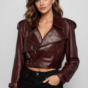 May include: A burgundy leather jacket with a notched collar, shoulder epaulets, and a belted waist. The cropped jacket features a single-button closure and long sleeves with buckle details. The model is wearing black jeans.