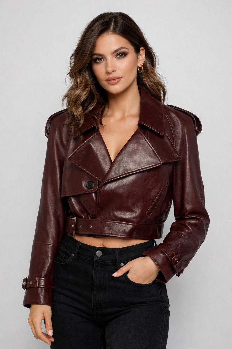 May include: A burgundy leather jacket with a notched collar, shoulder epaulets, and a belted waist. The cropped jacket features a single-button closure and long sleeves with buckle details. The model is wearing black jeans.
