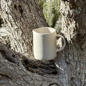 May include: A cream-coloured ceramic jug with a handle, set against a textured tree trunk. The jug has a simple, minimalist design, ideal for serving beverages. The background features soft greenery, suggesting an outdoor setting.