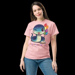 Puede incluir: Camiseta rosa claro con un diseño de serpiente de dibujos animados azul y turquesa, sosteniendo globos y una tarta de cumpleaños. El texto "HISS DAY QUEEN VIP" está impreso debajo. La camiseta la lleva una persona.