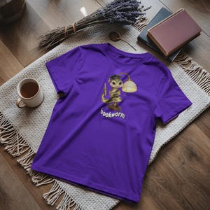 Può includere: Una maglietta viola con un disegno di un serpente dei cartoni animati con un cappello di laurea e avvolto attorno a una lampada. Il testo "bookworm" è sotto. La maglietta è su una superficie di legno con un tappeto decorativo e una tazza di tè.