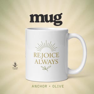 May include: White ceramic mug with the words "REJOICE ALWAYS" in gold lettering, a sunburst design, and a sprig of leaves. The mug also features the text "ANCHOR + OLIVE". The word "mug" is at the top.