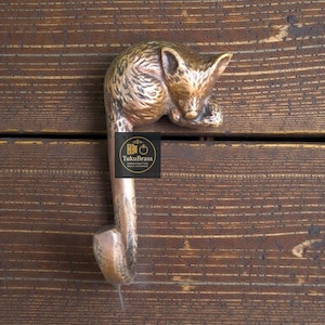 May include: A bronze-coloured metal hook in the shape of a sleeping fox. The hook is mounted on a wooden surface. The hook has a curved design with a small, rectangular end. The fox's fur is detailed with textured lines.