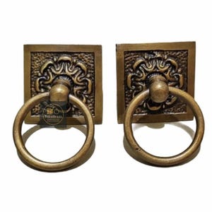 May include: Two antique brass door pulls. Each pull has a square backplate with an ornate floral design and a circular ring handle. The pulls are made of brass and have a warm, aged finish. They are designed for doors or drawers.