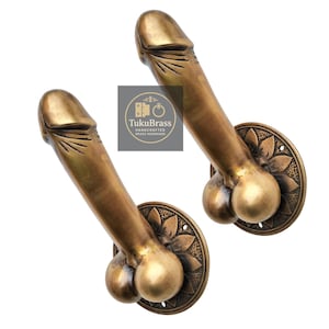 Puede incluir: Dos manijas de puerta de latón con diseño fálico. Cada manija presenta una escultura detallada de un pene y testículos, montada sobre una placa redonda con un patrón floral. Las manijas son de color dorado antiguo cálido.