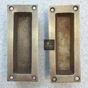 May include: Two rectangular brass recessed pulls with a worn, antique finish. Each pull has a long, recessed centre and four screw holes. The pulls are likely for a cabinet or drawer. The logo "TukuBrass" is visible.