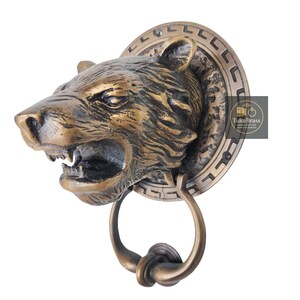 May include: A bronze-colored door knocker in the shape of a roaring tiger's head. The knocker features a circular backplate with a decorative border and a ring-shaped handle. The tiger's face has detailed features.