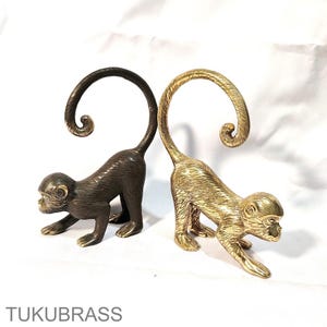 May include: Two brass monkey figurines. One is a dark bronze color, the other is gold. Both are in a crawling position with their tails curled upwards. The text "TUKUBRASS" is at the bottom.