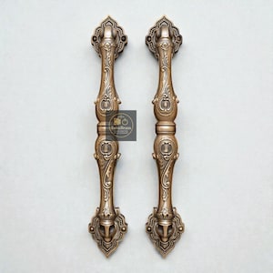May include: Two ornate bronze door handles with intricate floral and scrollwork designs. Each handle features a long, vertical grip with decorative elements at the top and bottom. The handles are symmetrical and have a vintage aesthetic.