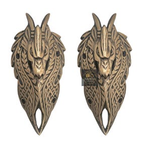 Könnte beinhalten: Zwei bronzefarbene dekorative Türgriffe. Jeder Griff zeigt ein stilisiertes Drachenkopf-Design mit komplizierten Knotenmustern und einer spitzen Basis. Die Griffe sind symmetrisch und scheinen aus Metall zu sein.