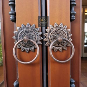 Könnte beinhalten: Nahaufnahme von zwei bronzefarbenen Türgriffen. Jeder Griff hat ein Sonnenblumen-Design mit einem Ringgriff. Die Griffe sind an einer Holztür mit braunem Finish angebracht. Der Markenname "TukuBrass" ist sichtbar.