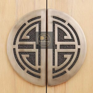May include: Two antique brass cabinet pulls with a circular design. Each pull is a half-circle with a textured, dark gray pattern inside the design. The pulls are mounted on a light wood cabinet door. The brand name "FukuBrass" is visible.