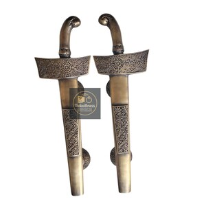 May include: Two antique brass door handles with ornate floral patterns. The handles have curved tops and detailed designs on the blades and grips. The handles are from TukuBrass, handcrafted.