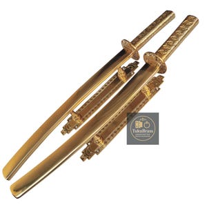 Puede incluir: Dos espadas katana decorativas de color dorado. Las espadas tienen empuñaduras y vainas detalladas. La imagen incluye el logotipo de TukuBrass Handcrafted Brass Hardware.