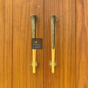 Puede incluir: Dos tiradores de armario de latón dorado con un diseño decorativo. Los tiradores son de color dorado con un patrón detallado y están montados en un armario de madera. Tienen una forma larga y delgada con una base circular y una parte superior decorativa.