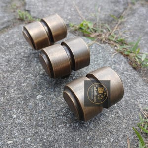 Peut inclure: Trois boutons de meuble en laiton antique avec un design cylindrique et une finition patinée. Les boutons sont disposés sur une surface en pierre grise, suggérant une esthétique rustique ou vintage. Les boutons ont une forme géométrique unique.