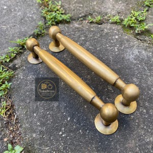 May include: Two antique brass door pulls with a cylindrical handle and ball-shaped ends. The pulls are a warm gold color and rest on a concrete surface. The logo "TukuBrass Handcrafted Hardware" is visible.
