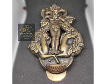 May include: A bronze-colored brass hardware piece featuring a sculpted figure with a trident, flanked by two winged horses. The design includes a serpent and is set against a dark background. The logo "TukuBrass" is visible.