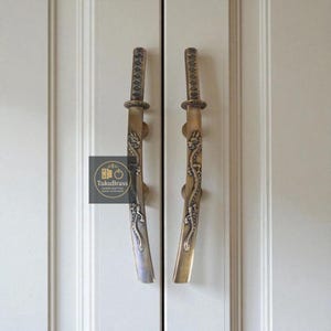 May include: Two bronze-toned cabinet handles shaped like Japanese katanas. The handles feature intricate dragon designs and are mounted on a white cabinet door. The handles have a textured grip and a decorative tsuba.