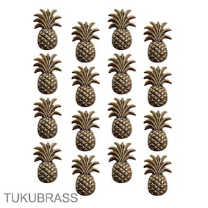 May include: Sixteen bronze pineapple-shaped cabinet knobs arranged in a grid. Each knob has detailed textures and a stylized pineapple design. The text "TUKUBRASS" is at the bottom left.