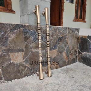 May include: Two brass door handles with a gold finish. The handles are long and cylindrical, with decorative bands and a flared base. The text "TUKUBRASS" is visible on the stone wall behind the handles.