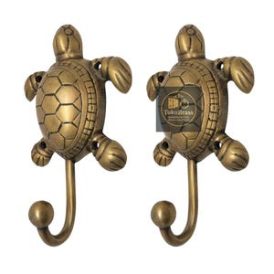 May include: Two brass turtle-shaped wall hooks. Each hook features a turtle design with a textured shell and curved hook. The hooks are made of brass and have a warm, golden color. The hooks are designed for hanging items.