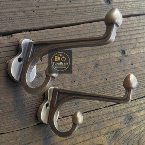 Op de afbeelding: Twee antieke messing kapstokken met een gebogen ontwerp en een afgerond uiteinde. De haken zijn gemonteerd op een houten oppervlak. Het logo "TukuBrass HANDCRAFTED BRASS HARDWARE" is zichtbaar.