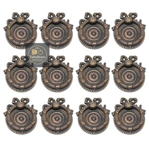 May include: Twelve antique bronze cabinet pulls with a floral centre design and a decorative bow at the top. Each pull has a ring handle. The pulls are arranged in a grid pattern.