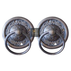 May include: Two antique-style bronze door pulls. Each pull features a circular backplate with an ornate floral design and a rounded handle. The pulls are made of metal and have a dark, aged finish. The pulls are designed for use on doors or drawers.
