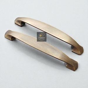 Könnte beinhalten: Zwei antike Messing-Schrankgriffe mit geschwungenem Design. Die Griffe haben eine rechteckige Basis und einen leicht gebogenen Griff. Die Griffe sind aus Messing und haben eine warme, goldene Farbe. Das Logo "TukuBrass" ist sichtbar.