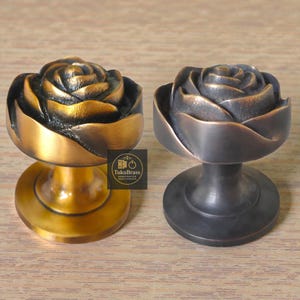 Puede incluir: Dos tiradores de armario con forma de rosa. Uno es dorado, el otro es bronce oscuro. Ambos tienen un diseño floral detallado y una base redonda. Los tiradores son de metal y son adecuados para muebles o decoración.