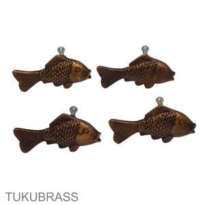 May include: Four bronze-colored fish-shaped cabinet knobs with silver screws. The knobs have detailed scale patterns and are arranged in a square formation. The text "TUKUBRASS" is at the bottom.