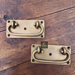 Puede incluir: Dos tiradores de cajón rectangulares de latón con acabado cepillado, cada uno con un asa central abatible unida a una placa trasera plana con cuatro orificios para tornillos en las esquinas y un orificio central. Logo TukuBrass.