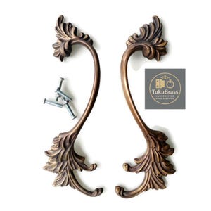 May include: A pair of antique bronze door handles with an elaborate leaf design. The handles are curved and feature decorative flourishes. Includes silver screws for installation. The logo "TukuBrass Handcrafted Brass Hardware" is visible.