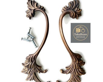 May include: Two antique bronze-toned metal cabinet handles featuring an ornate, curved leaf-like design at both ends. The set includes four silver-colored metal mounting screws. A square logo with the text TukuBrass Handcrafted Brass Hardware.