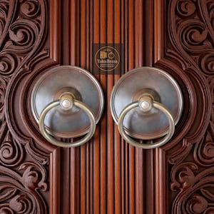 Può includere: Primo piano di una porta in legno marrone scuro con due battenti in ottone antico. I battenti hanno un design circolare con una maniglia ad anello. La porta presenta motivi floreali intagliati e venature del legno verticali.