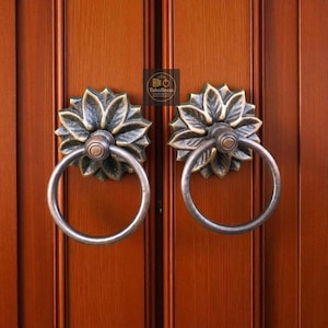 May include: A pair of antique door knockers with a floral design. Each knocker has a circular handle and a decorative base with a flower-like pattern. The knockers are made of metal and have a bronze finish. The door is made of wood.