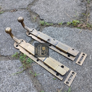 以下が含まれることがあります： 球状のハンドルが付いた、2つのアンティーク真鍮製のドアラッチ。ラッチは長方形で装飾的なエッジがあり、一致するストライクプレートが含まれています。画像には「TukuBrass HAND CRAFTED」というテキストが含まれています。