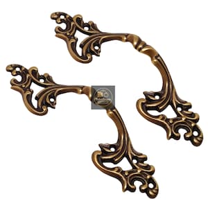 May include: Two ornate brass handles with a vintage aesthetic. The handles feature intricate floral and scrollwork designs, with a warm, aged finish. The handles are likely for furniture or cabinetry.