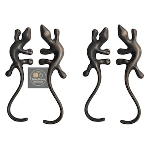 Könnte beinhalten: Vier bronzefarbene Schrankgriffe in Form von Eidechsen. Jeder Griff hat ein detailliertes Eidechsendesign mit einem gebogenen Schwanz. Die Griffe sind paarweise angeordnet, wobei sich die Schwänze in entgegengesetzte Richtungen krümmen. Das Logo "TukuBrass" ist sichtbar.